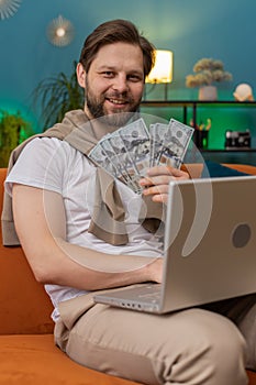 Smiling young man counting money cash and use laptop pc calculate domestic income earnings at home