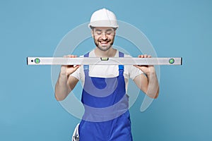 Smiling young man in coveralls protective helmet hardhat hold building bubble spirit level isolated on blue wall