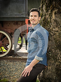 Smiling young man in denim shirt against tree near old train