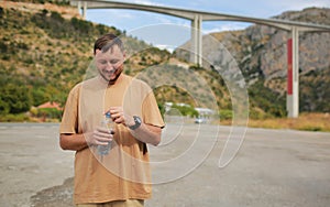Smiling young man drinking fresh water from plastic bottle outdoors after workout or running on hot summer day. Male
