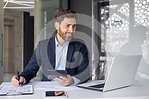 Smiling young man financial expert, businessman sitting in the office at the desk, working on a laptop and with