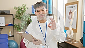 A smiling young man gesturing \'ok\' with clipboard in a physiotherapy clinic interior