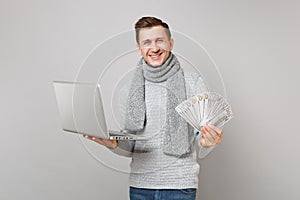 Smiling young man in gray sweater, scarf holding lots bunch of dollars banknotes cash money, laptop pc computer isolated