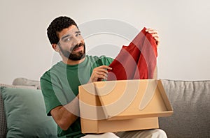 Smiling young man in a green shirt skeptically examines a red sweater just unpacked