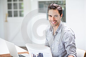 Smiling young man holding documents while sitting at desk with laptop