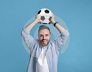 Smiling young man holding soccer ball above his head on blue studio background