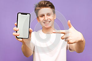Smiling young man holds a modern smartphone in his hand and shows his finger on a blank white screen, isolated on a blue