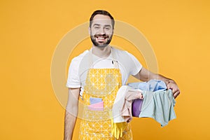 Smiling young man househusband in apron hold basket with clean clothes while doing housework isolated on yellow