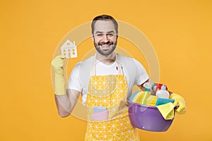 Smiling young man househusband in apron rubber gloves hold basin with detergent bottles washing cleansers doing