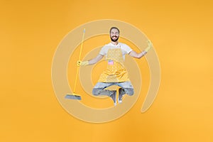 Smiling young man househusband in apron rubber gloves hold broom while doing housework isolated on yellow background