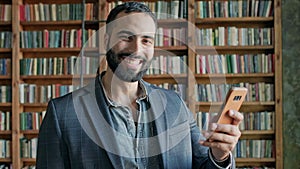 Smiling Young Man In The Library With Mobile Phone In His Hands. Stylish Guy With Beard In Jacket. He Gestured Okay.