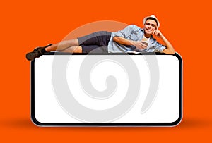 Smiling Young Man Lying On Big Blank Smartphone And Drinking Exotic Cocktail