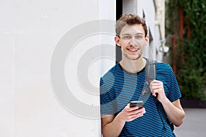 Smiling young man with mobile phone and backpack leaning against wall