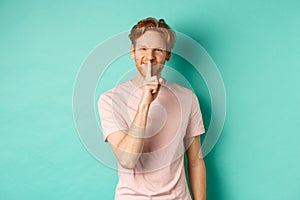 Smiling young man with red hair and beard sharing a secret, showing taboo gesture and grinning, shushing you to be quiet