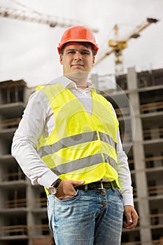 Smiling young man in red hardhat and green safety vest standing on building site