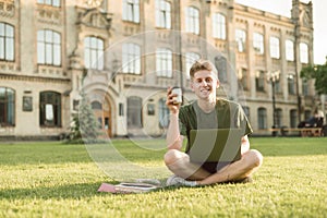 Smiling young man sits on a lawn on a university campus with a laptop on his lap, holds a cup of coffee in his hands, looks into