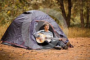 Smiling young man sitting near touristic tent and playing guitar