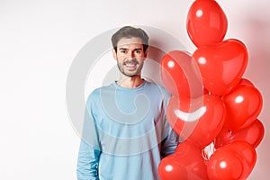 Smiling young man standing with heart balloons and looking happy, celebrating valentines day, bring romantic present to