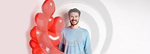 Smiling young man standing with heart balloons and looking happy, celebrating valentines day, bring romantic present to