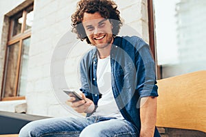 Smiling young man stitting outdoors texting on mobile phone. Happy male with curly hair using smart phone application for