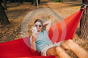 Smiling young man in sunglasses and casual clothes swings in a hammock in the woods at sunset and uses a smartphone with a happy