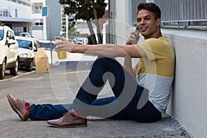 Smiling young man talking on smart phone while sitting on sidewalk