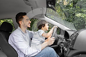 Smiling young man teaching little child to drive car in summer, boy hold steering wheel and look at road, profile