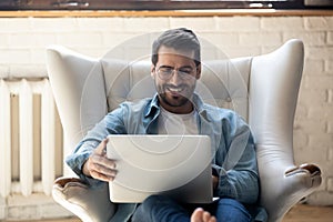 Smiling young man using laptop, sitting in cozy armchair