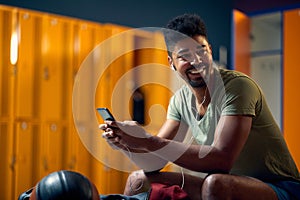 Smiling young man using smatphone in gym dressing room before workout