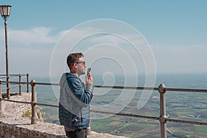 Smiling young man with wearing casual clothes over beautiful landscape background sending voice message on her smart phone.