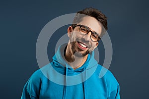 Smiling young man wearing glasses and bright hoodie standing against blue background