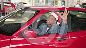 Smiling young man wearing red Christmas Santa hat and driving car, showing ok sign. New Year, celebration, holiday