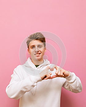 Smiling young man in white hoodie isolated on pink background, looks into camera with smile on face and shows thumbs up gesture of