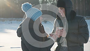 Smiling young man and woman talking standing with tea mugs in sunshine on snowy meadow at background on winter forest