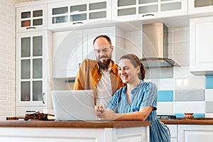 Smiling young married couple talking via video call using laptop at home in kitchen interior. Woman and man happy to
