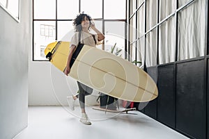 Smiling young mixed race woman arrives home listening to music and carrying a surf board after a day of surfing.