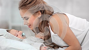 Smiling young mother admiring newborn baby kissing during waking up at white bedroom interior