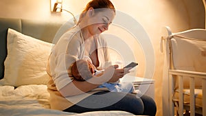 Smiling young mother feeding her newborn baby with breast milk working and browsing internet on smartphone at night in