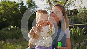 A smiling young mother hugs her little daughter, and blows soap bubbles. Summer park in the background. Slow motion