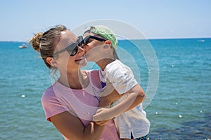 Smiling young mother kisses baby near the sea. Happy summer days.