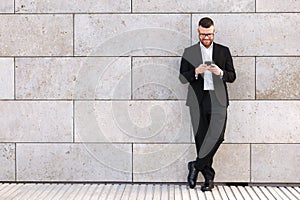 Smiling young office worker in formal suit standing outdoor with mobile phone in hands