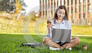 Smiling young professional woman multitasking with a laptop on her lap, enjoying a sandwich