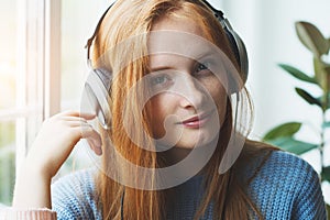 smiling young red haired girl with headphones listening to music at home, relaxing, enjoying songs