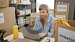 Smiling young redhead man enjoying volunteering at a charity center, sitting at a table with his laptop and headphones on