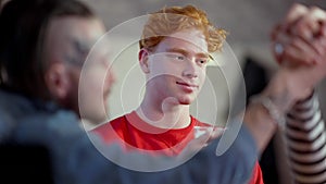 Smiling young redhead man looking at friends gambling armwrestling in pub. Cheerful Caucasian millennials having fun