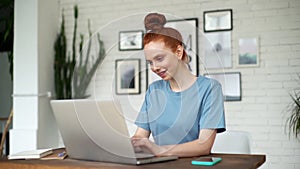 Smiling young redhead woman is typing on laptop at the desk at the home office.