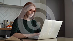 Smiling young redhead woman typing online social media message using laptop computer sitting at table in kitchen room