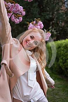 Smiling young redhead woman wearing stylish outfit near blossoming sakura in park. Fashionable spring look. Vertical frame