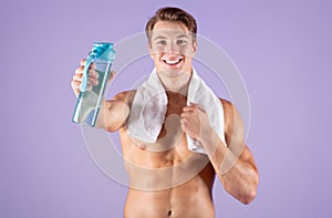 Smiling young sportsman with bare chest and towel showing bottle with fresh water on lilac studio background