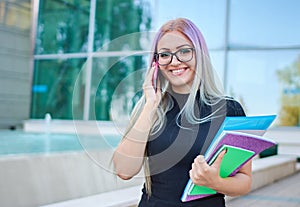 Smiling young student girl from z generation standing in a campus in front of a university building facade and talking on